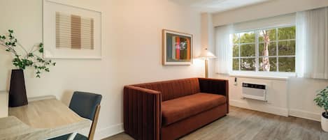 A modern living room with a brown sofa, a wooden coffee table, and a chair. There is a window with curtains, a wall-mounted lamp, and a framed picture.