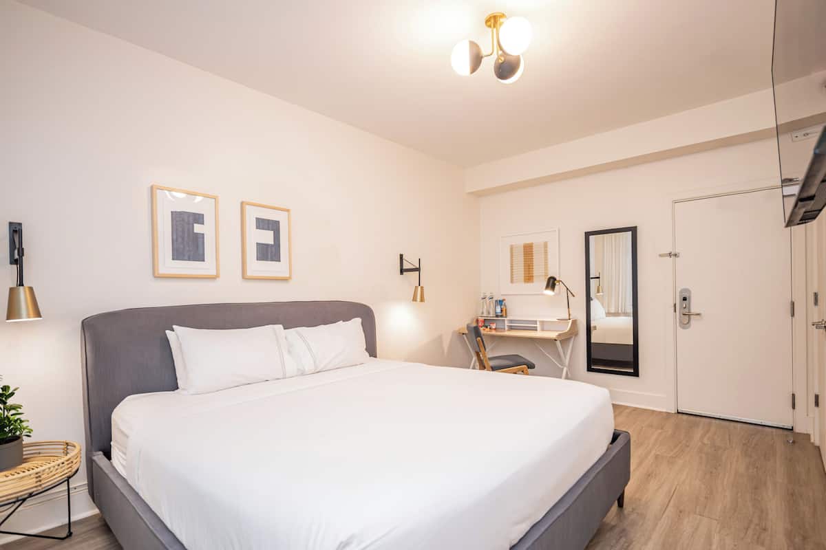 A modern hotel room with a large bed, bedside table, lamp, and a desk with a chair.