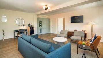 Una sala de estar moderna con un sofá azul, una mesa redonda de centro y dos sillones. Se aprecia una pequeña mesa con una lámpara y una planta. Al fondo, se distingue una zona de cocina.