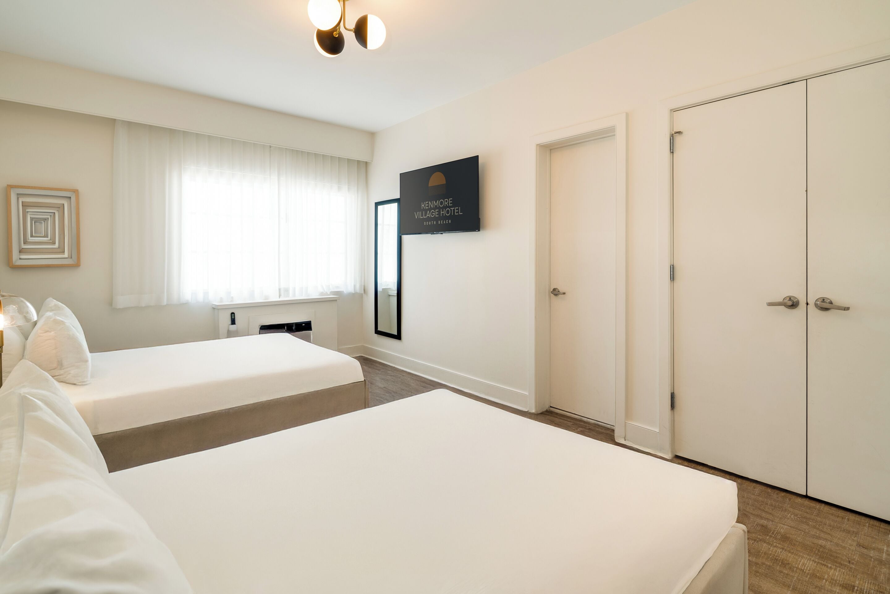 A hotel room with two beds, a large window with sheer curtains, a framed sign on the wall, and a wardrobe with a mirror.