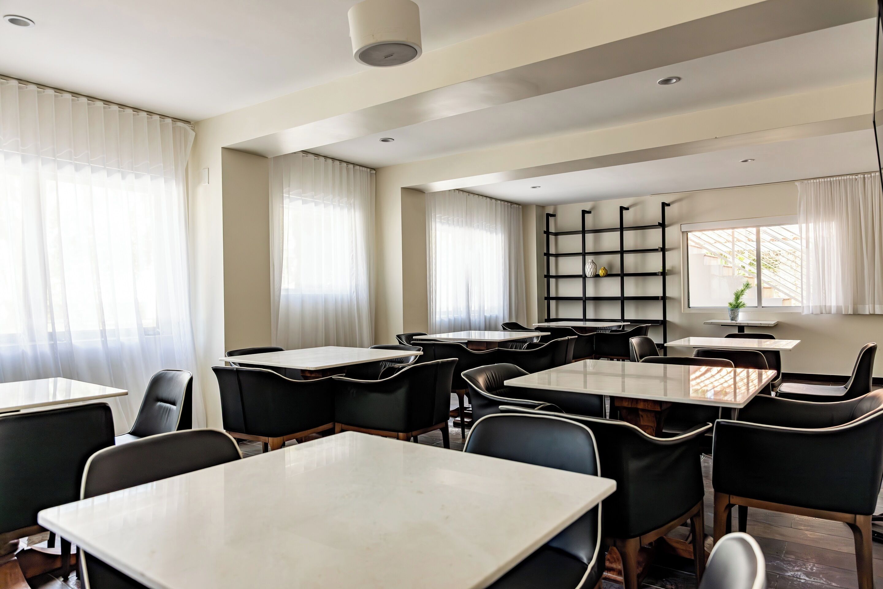 A modern dining area with tables and chairs, large windows with sheer curtains, and a black bookshelf.