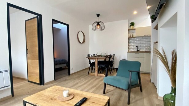 Una sala de estar moderna con una mesa de centro de madera, un sillón azul verdoso, una zona de comedor con una mesa y sillas de madera, una cocina con armarios blancos y una lámpara colgante.
