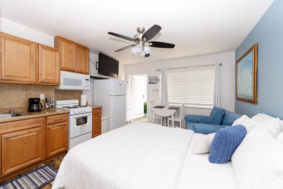 Quarto de hotel compacto com armários de madeira, cozinha compacta, cama com roupa de cama branca e azul, ventilador de teto e janela com persianas.