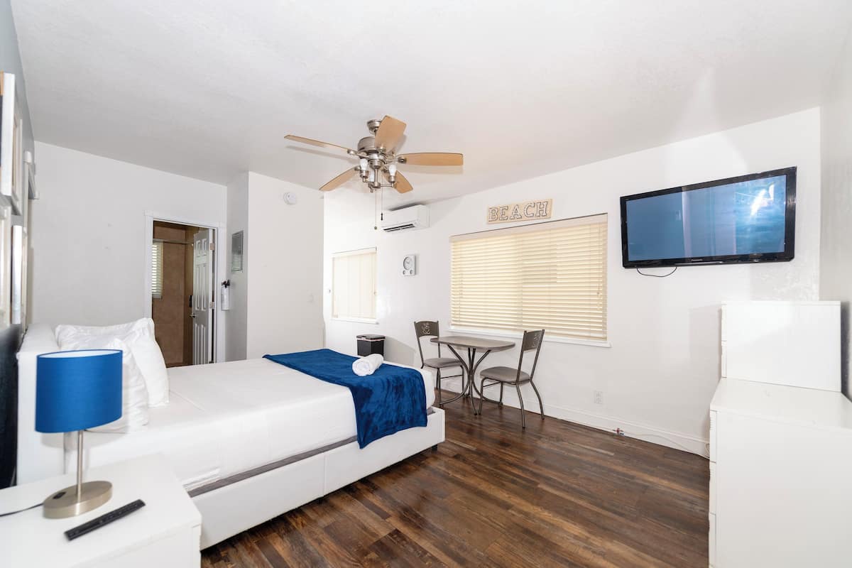 Quarto de hotel com cama, escrivaninha, cadeira, ventoinha no teto, televisão e janela com persianas.