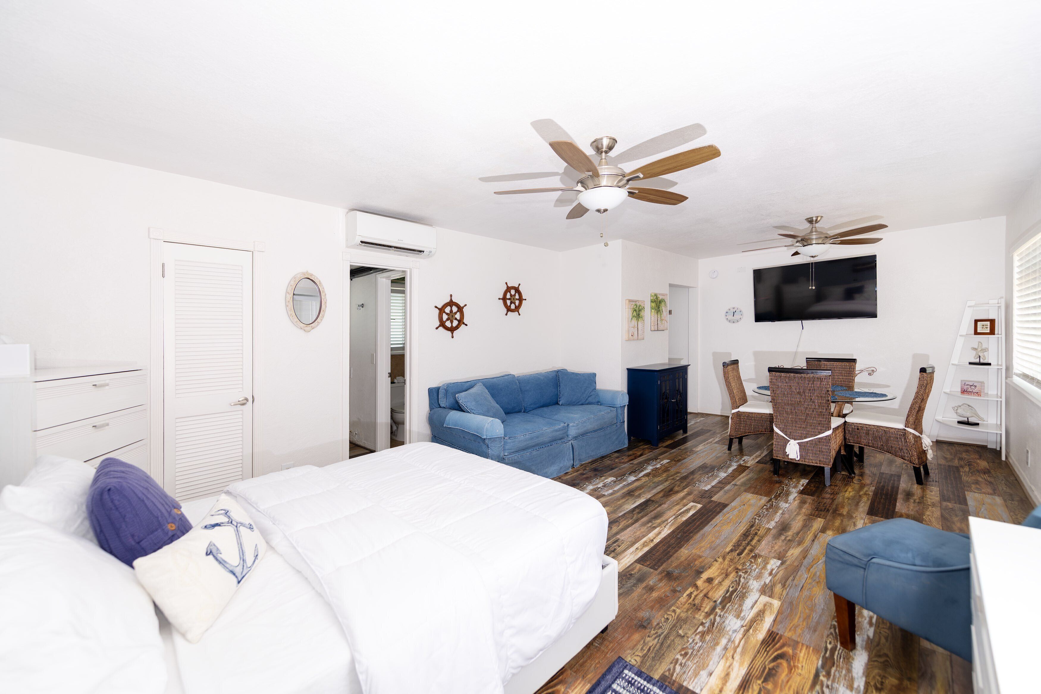 Une chambre à coucher avec un lit, un canapé bleu, une table à manger avec des chaises, un ventilateur de plafond et un téléviseur fixé au mur.