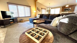 A living room with a sectional sofa, a TV, and a wooden XOXO game on a coffee table. - Lake Arrowhead Retreat with club pools and beaches (Nekoosa)