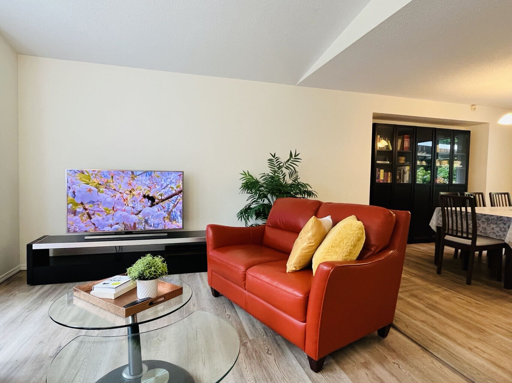 Ein Wohnzimmer mit rotem Sofa, einem Glastisch, einem Flachbildfernseher, einem Bücherregal und einem Essbereich.