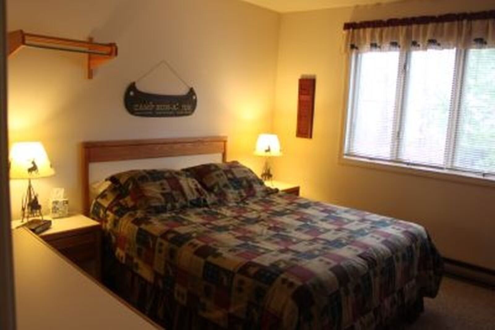 A neatly made bed with a quilt, two bedside lamps, a wooden headboard, a wall-mounted sign, and a window with curtains.