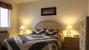Ein Schlafzimmer mit einem Bett, zwei Nachttischen mit Lampen, einer Kommode, einem Fenster mit Jalousien und einem gerahmten Bild an der Wand.