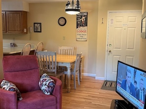 A living room with a red armchair, a wooden dining table with chairs, a television displaying a news channel, and a door with a sign.