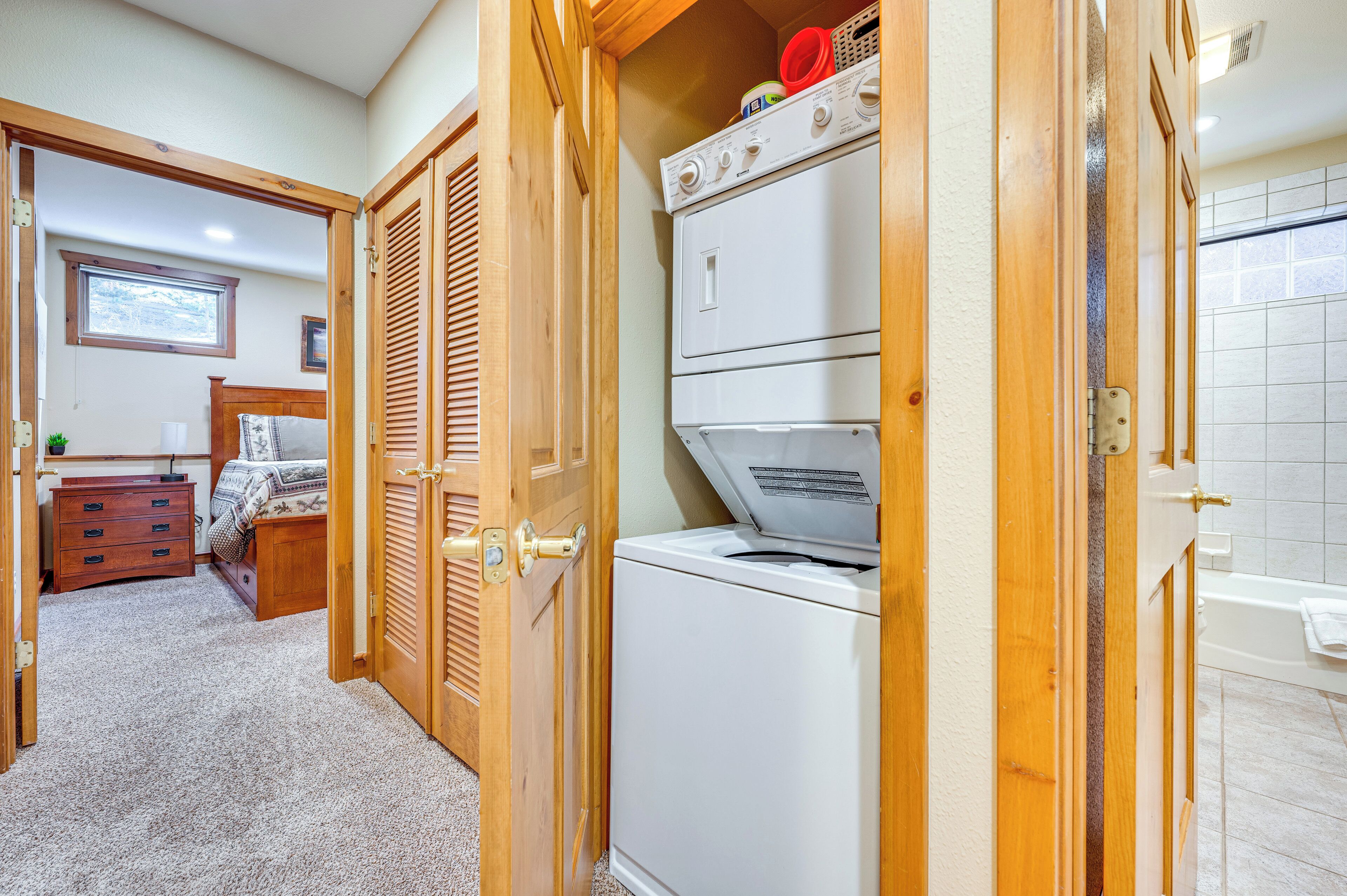 A laundry room with a stacked washer and dryer, wooden doors, and a view into a bedroom with a bed and dresser.