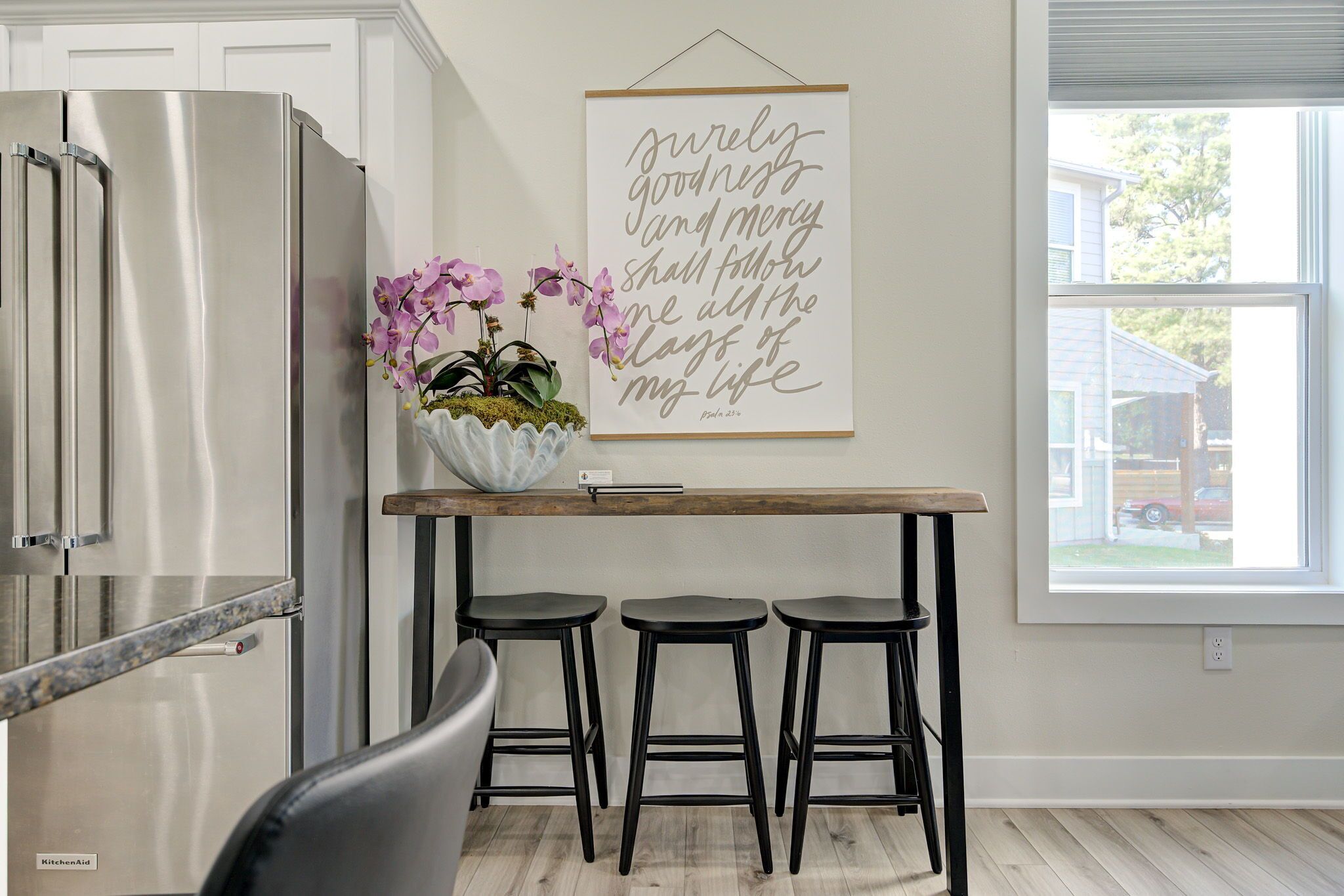 Una cocina con un refrigerador de acero inoxidable, una mesa de madera con tres taburetes negros y un letrero en la pared con un versículo bíblico.