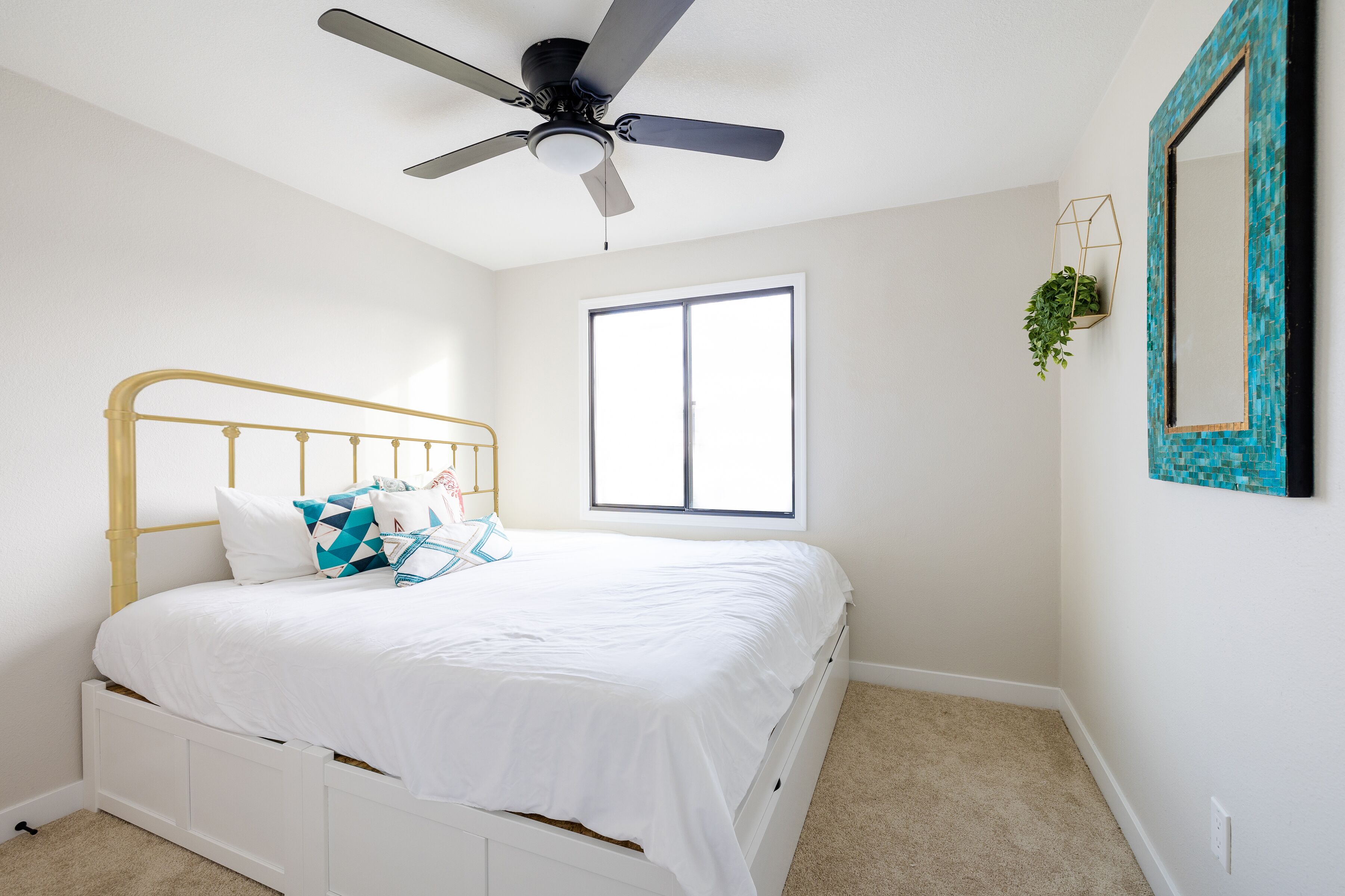 Un dormitorio con una cama blanca, un ventilador de techo, una ventana y un espejo montado en la pared.