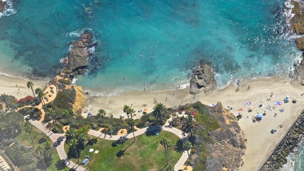 Une vue aérienne d’une plage aux eaux turquoise et limpides, parsemée de formations rocheuses et bordée d’une promenade bien entretenue, agrémentée de palmiers.