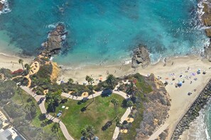 Luchtbeeld van een strand met helder turquoise water, rotsachtige kliffen en een verzorgde promenade met palmbomen.