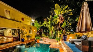 Une piscine dotée d’une terrasse carrelée, entourée de plantes en pot et bordée d’un bâtiment jaune avec une fenêtre.
