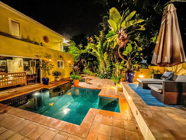 A swimming pool with a tiled deck, surrounded by potted plants and a yellow building with a window.