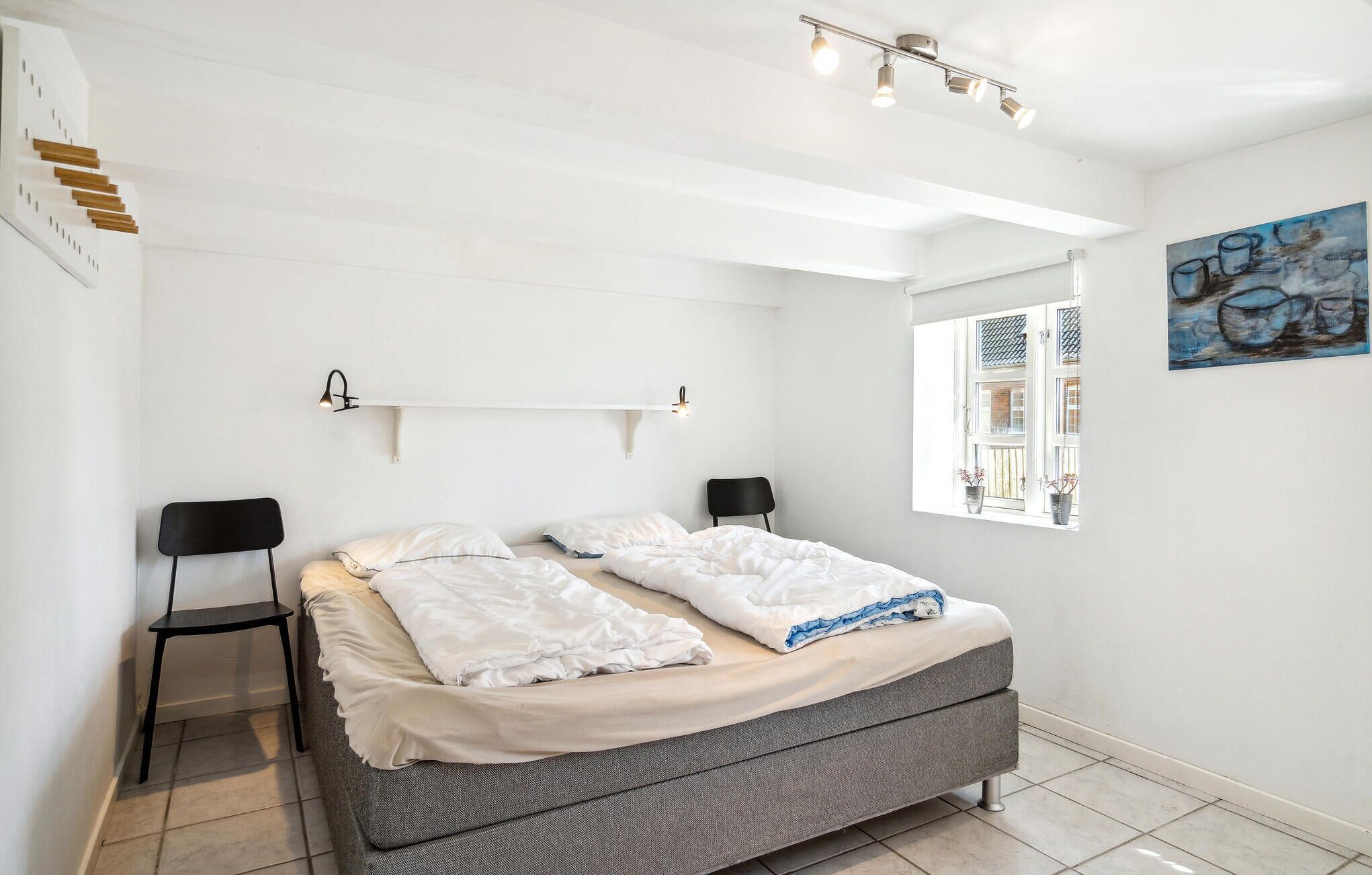 A bedroom with a bed, two chairs, a shelf, a painting, and a window with a view.