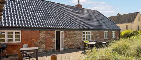 Une maison en briques avec un toit en tuiles, une terrasse avec des meubles d’extérieur et un ciel dégagé.