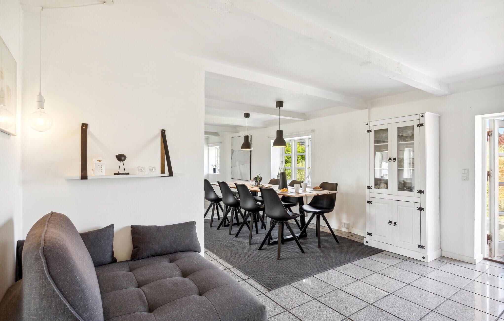 Un salon moderne avec un canapé gris, un coin repas avec une longue table et des chaises, et un meuble blanc avec des portes en verre.