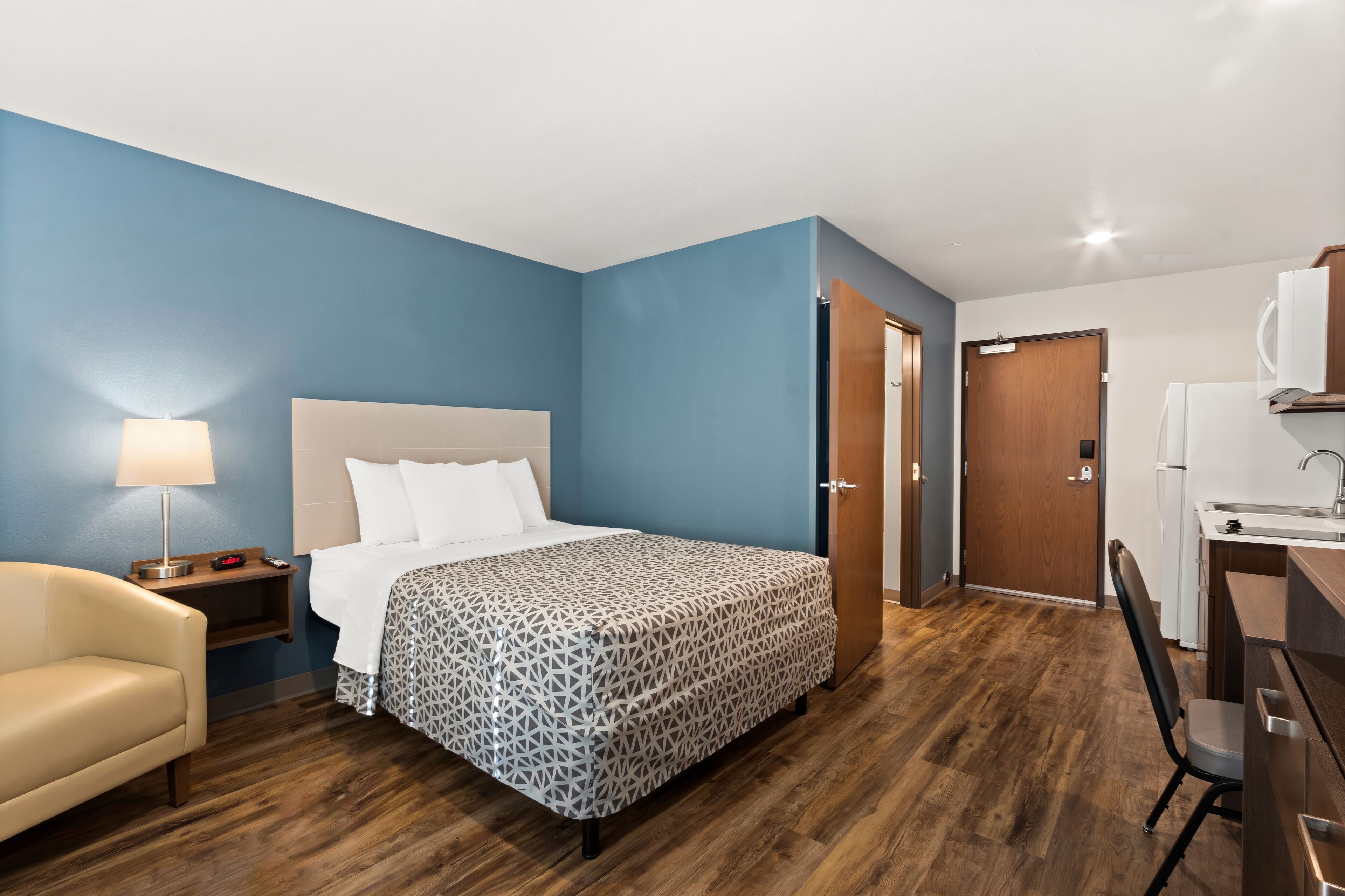 Una habitación de hotel con cama, mesita de noche, lámpara, silla, escritorio y kitchenette.