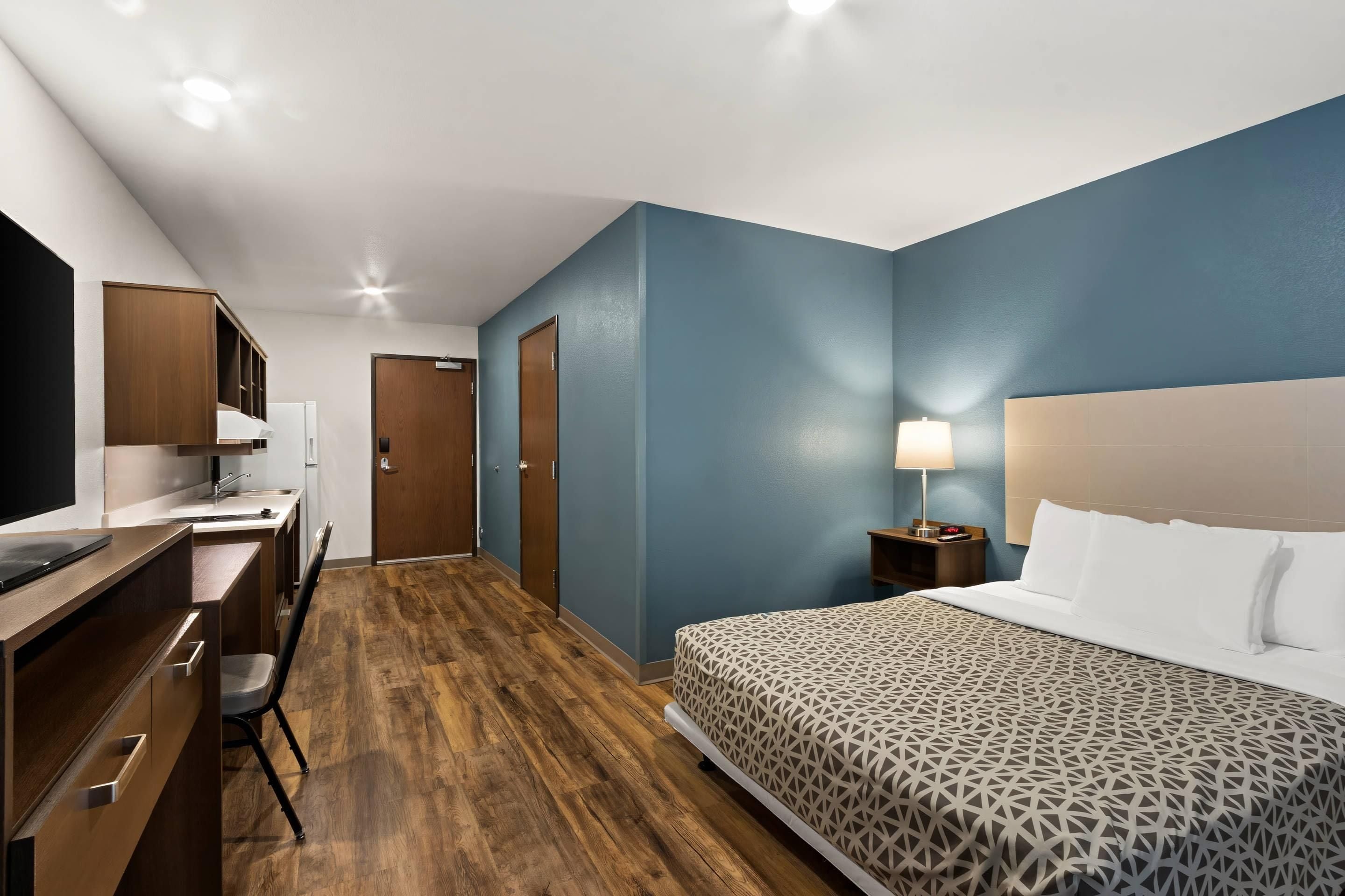 A hotel room with a bed, a desk with a chair, a nightstand with a lamp, and a bathroom door.