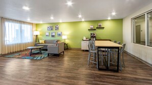 A modern living room with a sofa, coffee table, and dining table. There are framed pictures on the wall and a lamp on a side table. - Woodspring Suites Tolleson - Phoenix West (Tolleson)