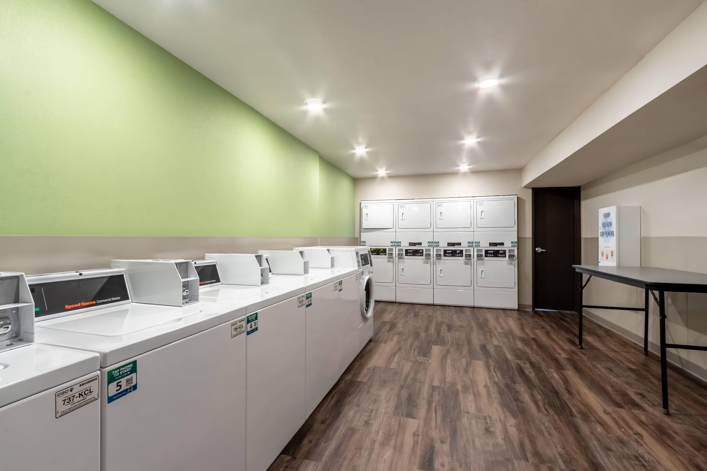 Una sala de lavandería con varias lavadoras y secadoras, suelo de madera y pared verde.