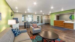 A modern living room with a sofa, armchair, and coffee table, a dining area with chairs and a table, and a kitchen area with a counter and cabinets. - Woodspring Suites Tolleson - Phoenix West (Tolleson)