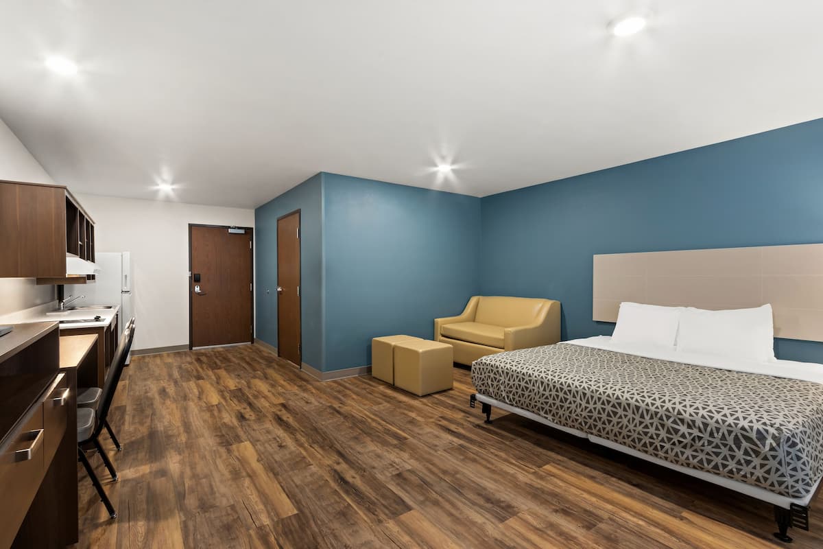 Ett hotellrum med trägolv, en säng med ett mönstrat överkast, en beige fåtölj, ett skrivbord med en stol och en garderob.