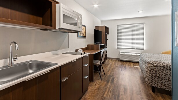 A compact kitchen with a microwave, sink, and wooden cabinets. It includes a small dining area with a chair and a bed with a patterned bedspread. - Woodspring Suites Tolleson - Phoenix West (Tolleson)