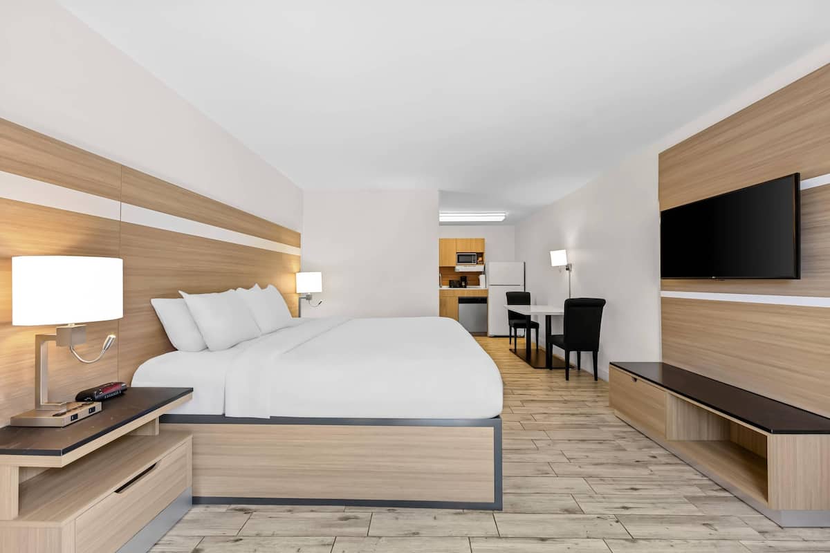 Ein modernes Hotelzimmer mit Bett, Nachttisch, Lampe, Fernseher und einer Küchenzeile im Hintergrund.