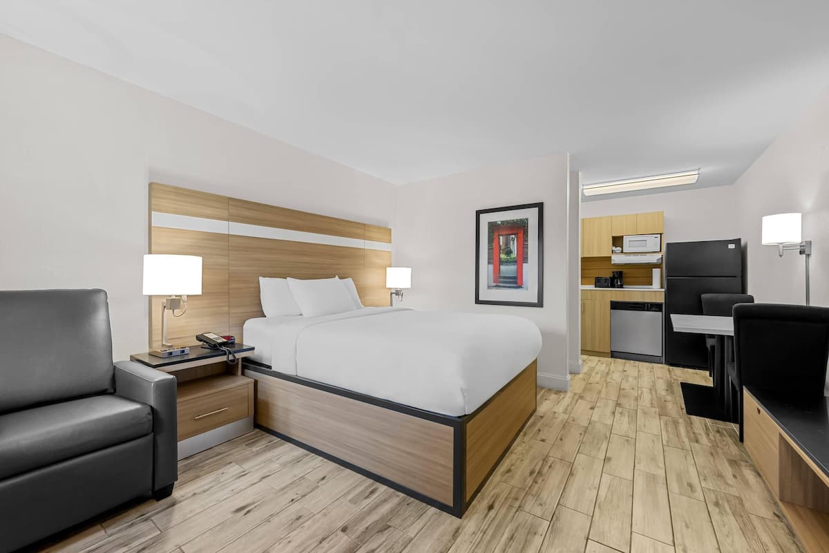 Ein Hotelzimmer mit Bett, Nachttisch, Lampe, Stuhl und einer Küchenzeile.