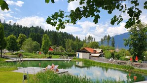 Un cadre paisible au bord d’un lac, avec une jetée en bois, des chaises longues et un petit bâtiment, le tout entouré d’une végétation luxuriante et dominé par une chaîne de montagnes.