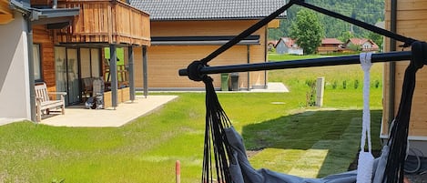 A wooden house with a balcony, a hanging chair, and a grassy yard with mountains in the background.