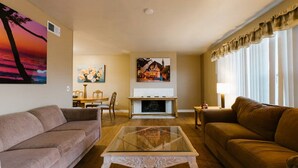 Una sala de estar con un sofá color beige, un sofá marrón, una mesa de centro de vidrio, una chimenea y un comedor con una mesa y sillas.