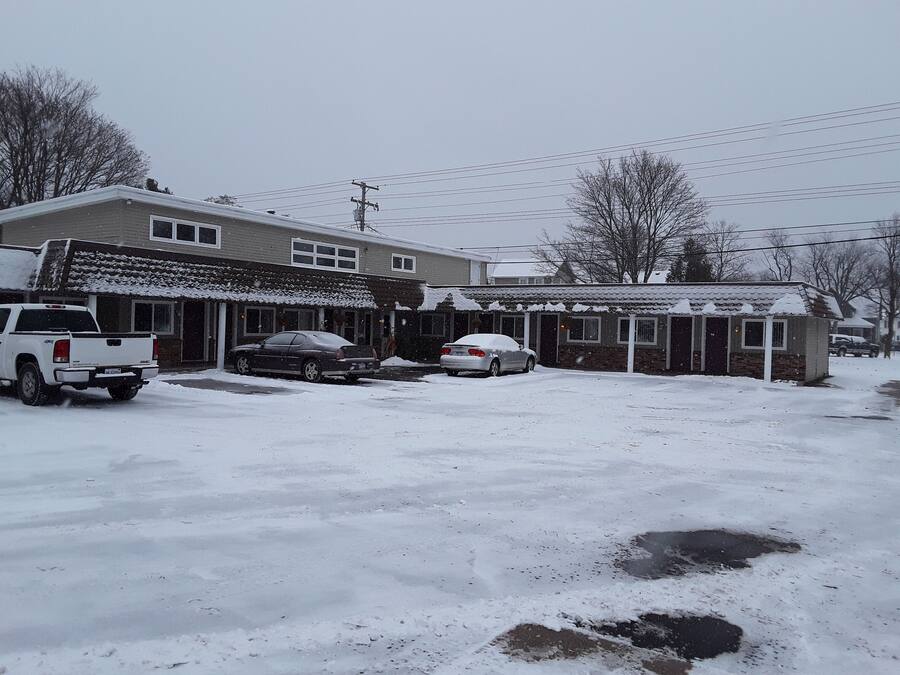 Zona residencial con una casa, un garaje y un cobertizo pequeño. Dos coches están estacionados frente a la casa, y el suelo está cubierto de nieve.