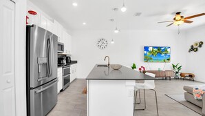 A modern kitchen with a stainless steel refrigerator, a microwave, and a sink. There is a dining area with a table and chairs, and a ceiling fan.