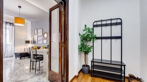 Une pièce avec un plancher en bois, un coin repas avec une table et des chaises, un présentoir en métal noir, une plante en pot et une porte donnant sur une autre pièce.