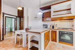 Une cuisine avec des meubles en bois, un plan de travail blanc, un coin repas avec une table et des chaises blanches, et une horloge au mur.