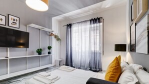 Une chambre d’hôtel moderne, équipée d’un lit, d’une télévision, d’une plante et d’une fenêtre avec des rideaux légers.