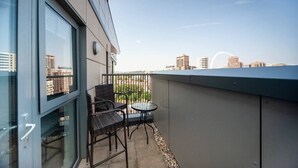 Un balcón con puerta de cristal, dos sillas de mimbre, una pequeña mesa redonda y vistas a edificios de la ciudad.
