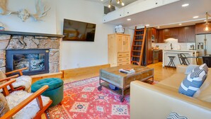 A modern living room with a stone fireplace, a wooden cabinet, and a kitchen area with wooden cabinets and a stainless steel refrigerator. - Copper Mtn Condo w/ Heated Pool: Ski-In/Walk-Out! (Frisco)