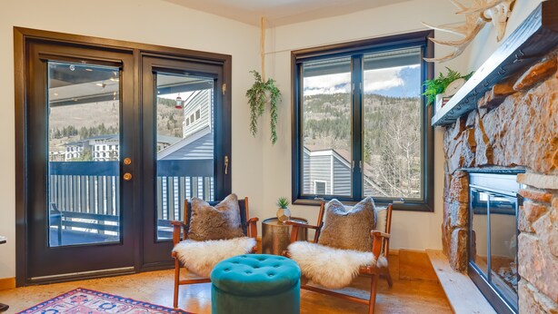 A cozy room with a fireplace, two chairs with fur throw pillows, a round ottoman, and a view of a wooden deck and mountains through large windows. - Copper Mtn Condo w/ Heated Pool: Ski-In/Walk-Out! (Frisco)