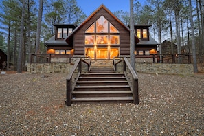 A log cabin with a stone foundation, wooden steps leading to a large front door, and a view of the surrounding forest. - Summer Sale Luxury Cabin! 4 Kings, sleeps 14. (Broken Bow)