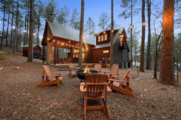 A cabin with a fire pit and Adirondack chairs in a forest setting. - Summer Sale Luxury Cabin! 4 Kings, sleeps 14. (Broken Bow)