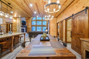 A wooden cabin interior with a large dining table, bar stools, a stone fireplace, and a TV mounted on the wall. - Summer Sale Luxury Cabin! 4 Kings, sleeps 14. (Broken Bow)