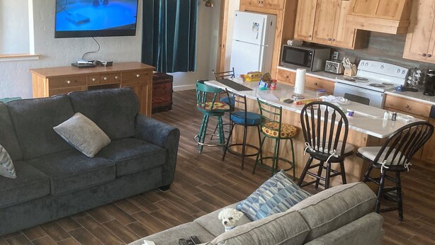 A modern living room with a grey sofa, a dining area with a table and chairs, wooden cabinets, a kitchen with a stove and microwave, and a TV on the wall. - Amazing views close to friends, family and fishing. Indoor RV parking 29’ max.  (Council)