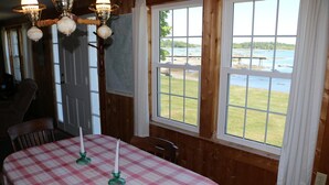 A dining area with a table set for two, featuring a checkered tablecloth and candles. There are two windows offering a view of a grassy area and a body of water. - Point Field (Deer Isle)
