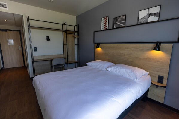 A modern hotel room with a large bed, a desk, and a chair. There are wall-mounted shelves with framed pictures and a small speaker on the wall.